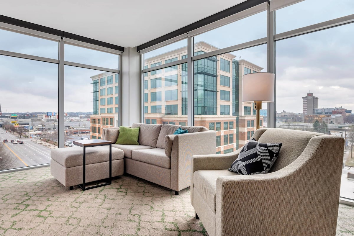 Un salon moderne avec un canapé d’angle, une petite table et une lampe sur pied. De grandes fenêtres offrent une vue sur le paysage urbain et sur un immeuble aux fenêtres en verre.