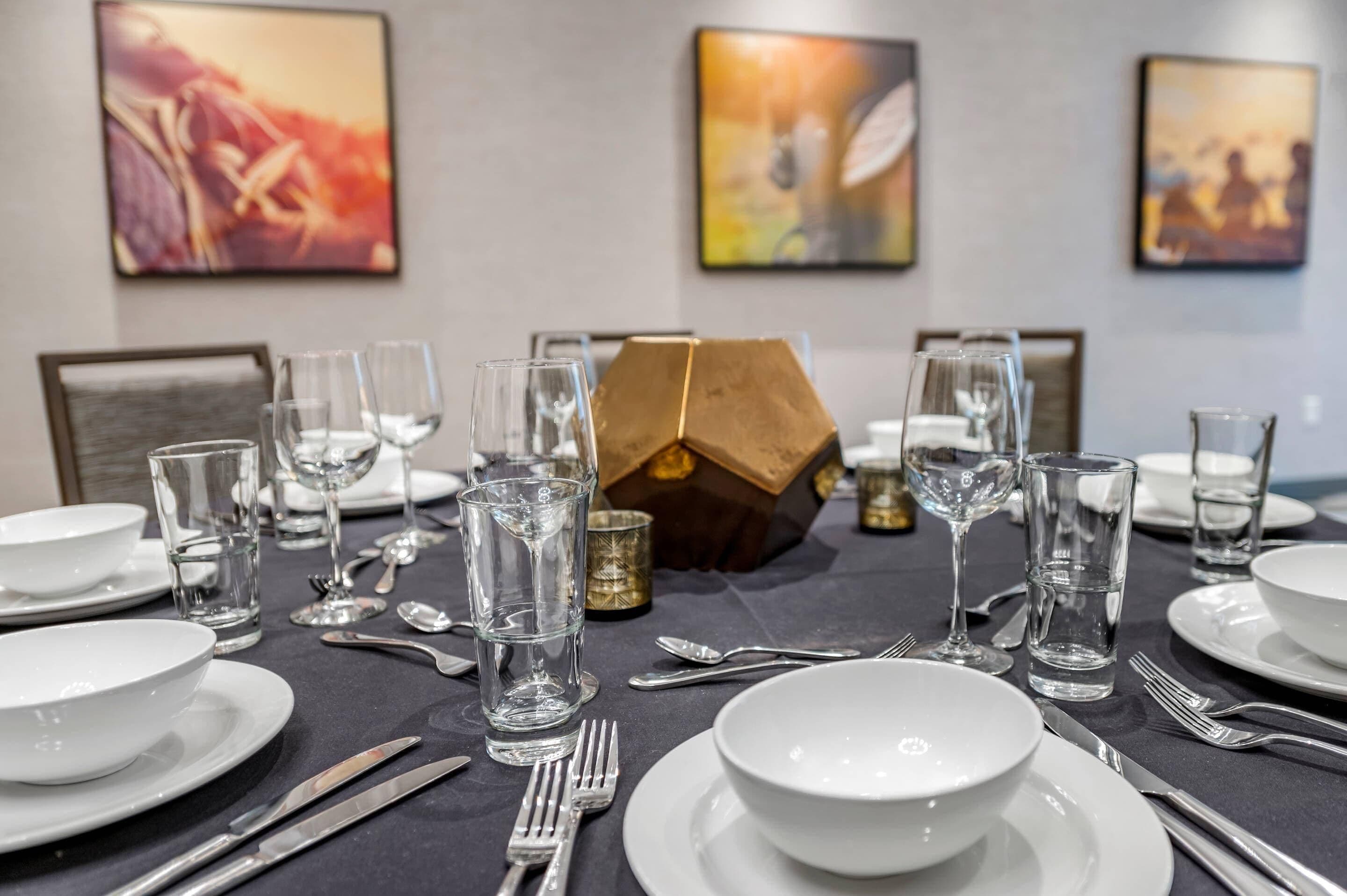 A dining table set with white plates, bowls, cutlery, and glasses, with abstract paintings on the wall behind.