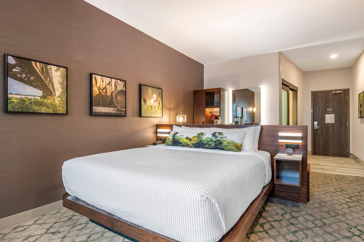 Une chambre d’hôtel avec un grand lit, une tête de lit en bois, une table de chevet et un miroir. Trois tableaux encadrés sont accrochés au mur.