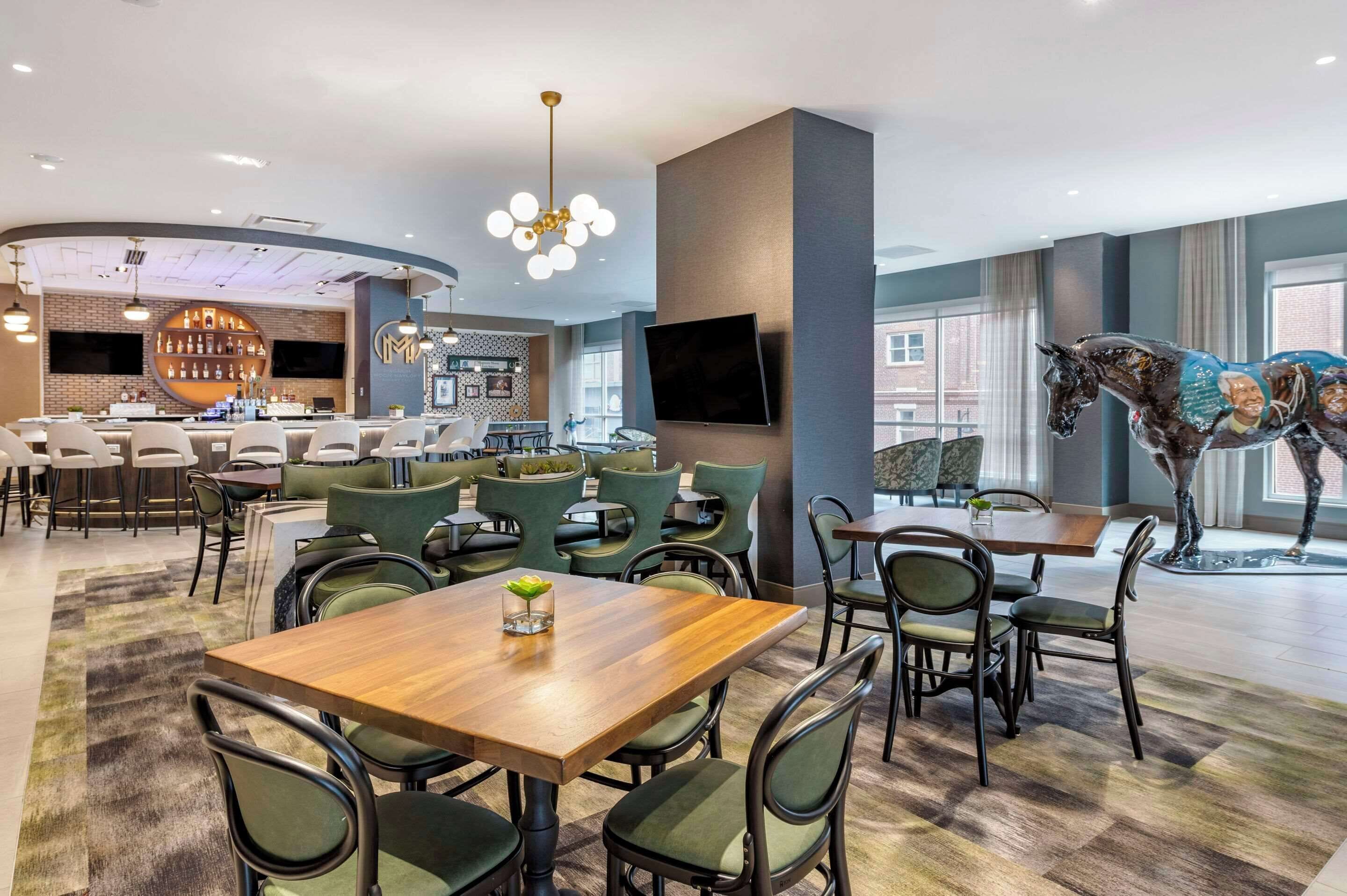 A modern dining area with a bar, green upholstered chairs, and a large horse statue.