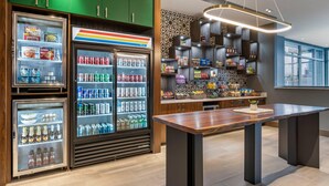A modern interior with a central wooden table, refrigerated beverage dispensers, and shelves stocked with snacks and condiments. - Cambria Hotel Louisville Downtown-Whiskey Row (Louisville)