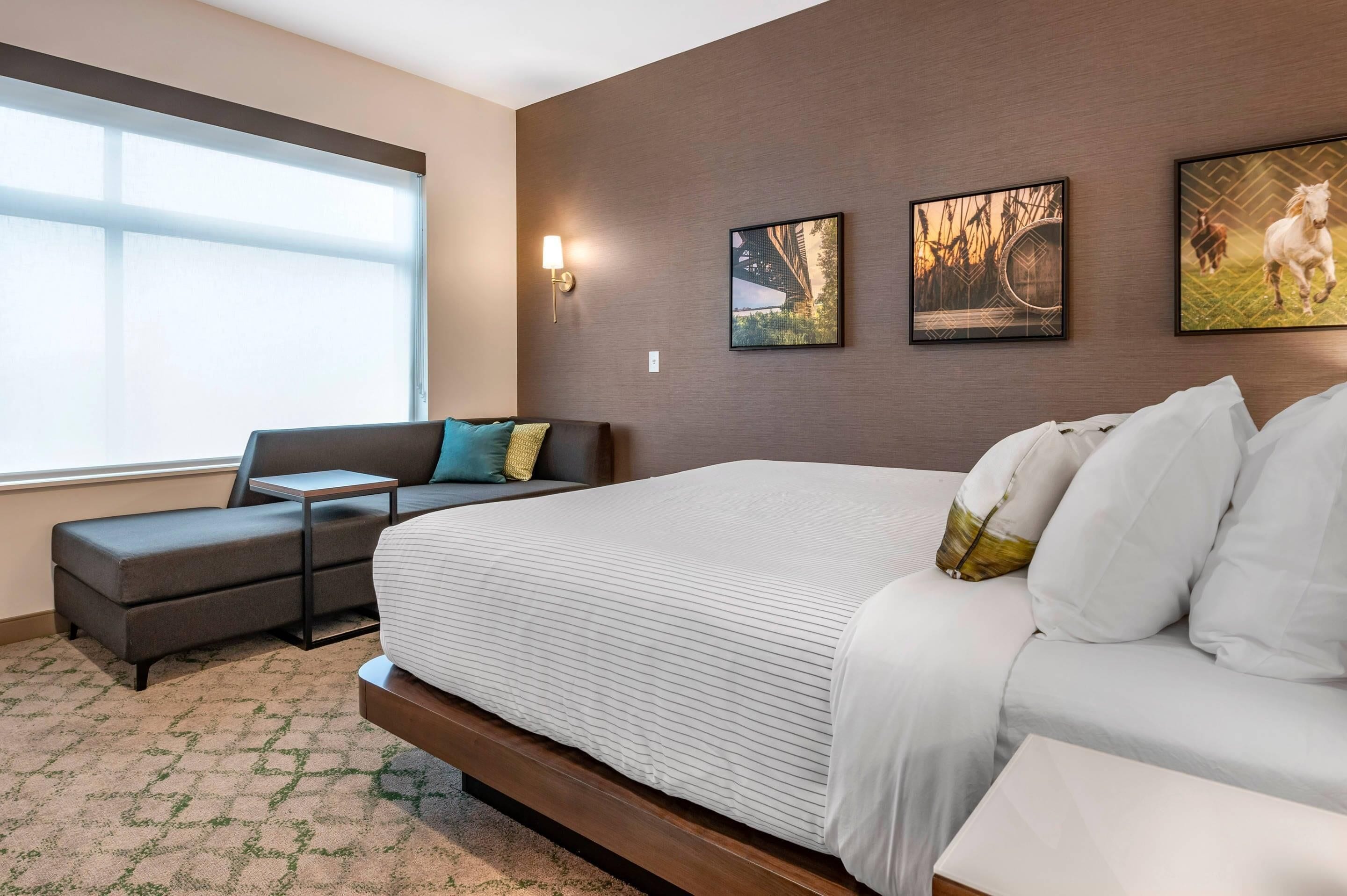 Quarto de hotel com uma cama grande, um sofá, uma mesinha e uma janela. Há três quadros na parede.