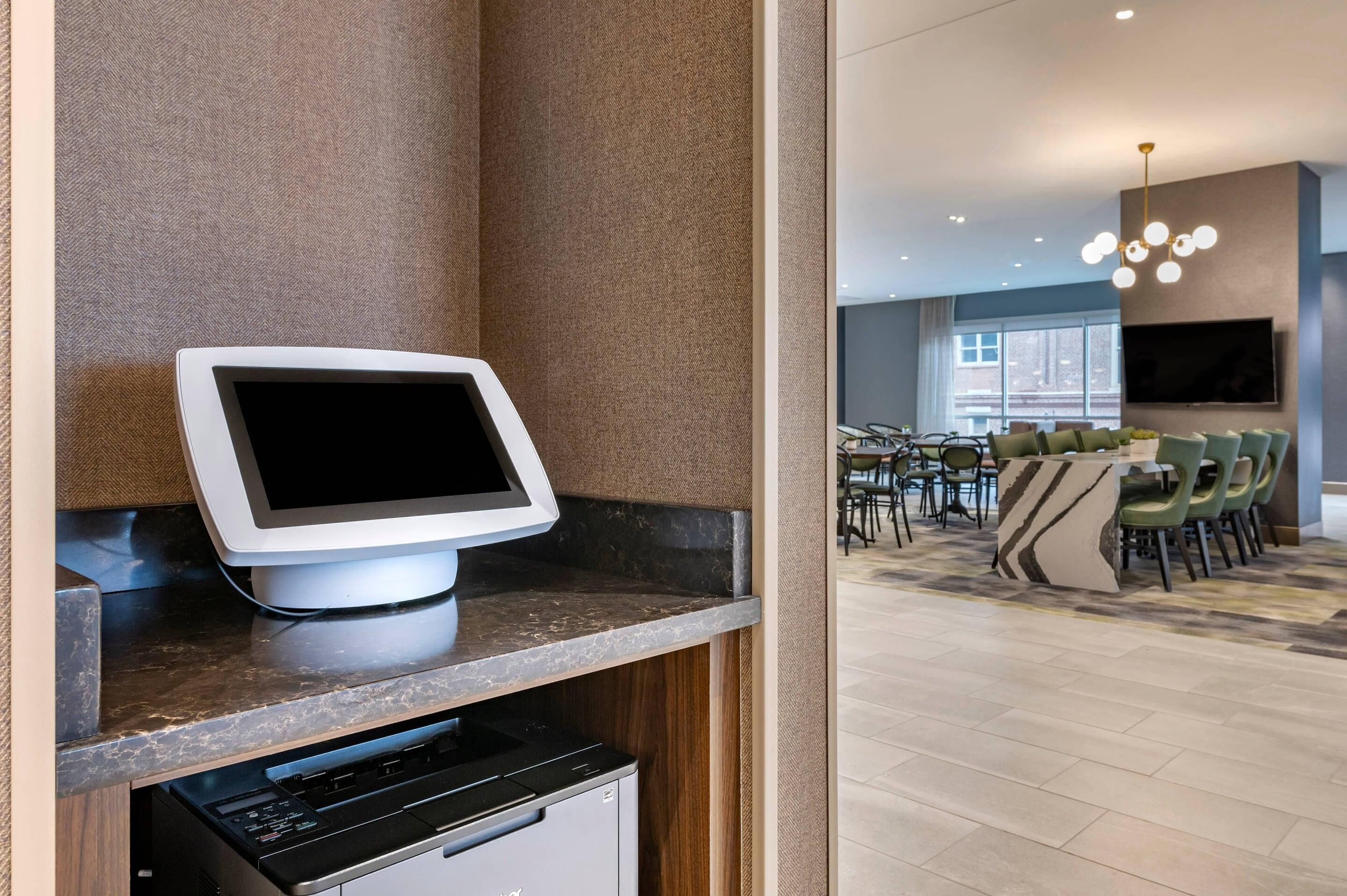 Una habitación de hotel moderna con un quiosco digital integrado, un área de comedor con sillas verdes y un televisor grande montado en la pared.
