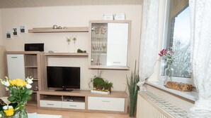 A modern living room with a flat-screen TV, wooden furniture, a dining table with a black rug, and a window with a view of trees. - 2 Ferienwohnungen in Seenähe (Bitterfeld-Wolfen)