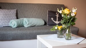 A living room with a grey sofa, a rolled-up light blue blanket, and a black pillow with an anchor design. There are two glass vases with yellow and white flowers on a white table. - 2 Ferienwohnungen in Seenähe (Bitterfeld-Wolfen)