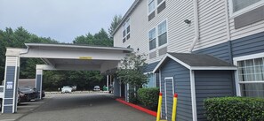 A building with a covered entrance, a sign indicating clearance, and a parking area with cars.