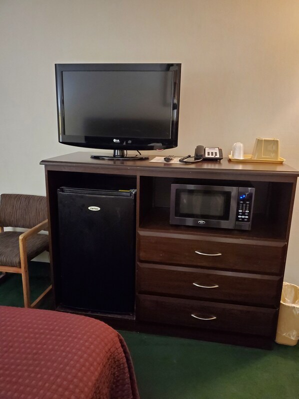 A compact hotel room with a flat-screen TV, a mini-fridge, a microwave, and a small table with a phone and a cup.