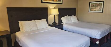 A hotel room with two beds, each with white linens and dark headboards. There is a nightstand with a lamp between the beds.