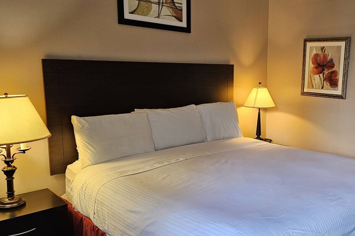 A neatly made bed with white linens, a dark headboard, a bedside lamp, a framed floral artwork on the wall, and a classic wooden chair.