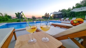 A poolside setting with two glasses of a yellow beverage, lounge chairs, a fruit bowl, and a view of the pool. - Amazing New Home in the Heart of Istria (Sveti Petar u Šumi)