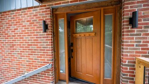 A wooden front door with a glass panel, set in a brick wall with a metal gutter and a wooden bench to the right. - The Church (Hendersonville)