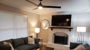 A living room with a fireplace, a sofa, a coffee table, and a TV mounted above the fireplace. - Renovated 3BR/2bath Ranch Near Hospital-Ideal for Families & Medical Stays (Merriam)