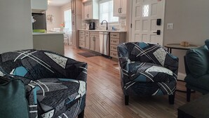 A modern living room with patterned armchairs and a wooden floor. A kitchen with white cabinets and a dining area with a table and chairs is visible in the background. - Renovated 3BR/2bath Ranch Near Hospital-Ideal for Families & Medical Stays (Merriam)