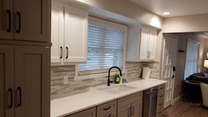 A modern kitchen with white cabinets, a tiled backsplash, a stainless steel sink, and a window with blinds. - Renovated 3BR/2bath Ranch Near Hospital-Ideal for Families & Medical Stays (Merriam)