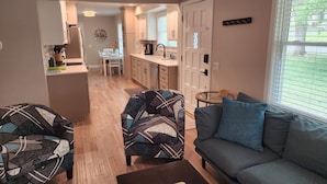 A living room with a grey sofa, patterned armchairs, and a coffee table. A kitchen with white cabinets and a dining area with a table and chairs is visible in the background. - Renovated 3BR/2bath Ranch Near Hospital-Ideal for Families & Medical Stays (Merriam)