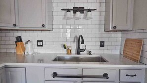 A kitchen with white cabinets, a stainless steel sink, and a black faucet. There are three light fixtures above the sink, and a wooden cutting board is visible on the countertop. - Housepitality - Top Choice Flat - Great Location (Columbus)
