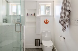 A modern bathroom with a glass shower enclosure, a white toilet, and a shelf with toiletries. - Modern cottage with full kitchen & private patio. Close to great hiking trails! (Oakland)