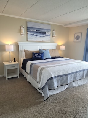 A neatly arranged bedroom with a bed, two bedside tables, a nightstand, a wall-mounted lamp, a framed picture, and a ceiling light fixture.