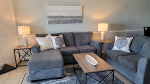 A modern living room with a grey sectional sofa, two side tables with lamps, a wooden coffee table, and a large abstract painting on the wall. - 2027 OPEN FOR BOOKING!  PIERVIEW 107 OCEAN-BEACHFRONT CONDO, 2BD&2BA SLEEPS 6 (Nags Head)