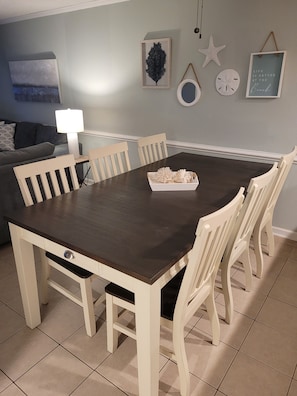 A dining area with a dark wooden table surrounded by white chairs, a small side table with a lamp, and decorative items on the wall.