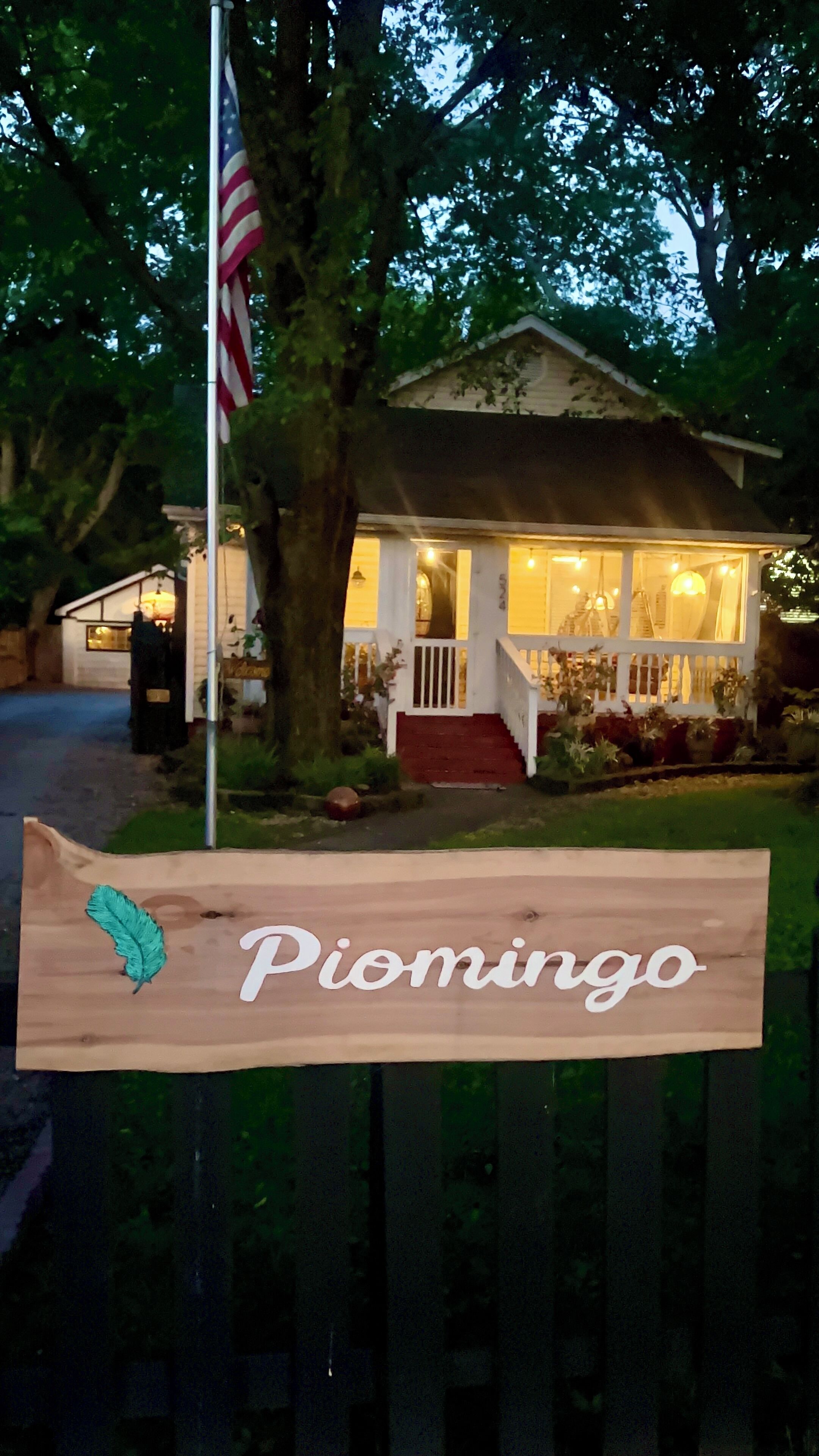 A wooden sign with the name "Piomingo" in front of a house with a porch, a flag, and a well-lit front yard.