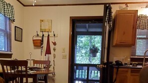 Un salon chaleureux avec un plancher en bois, un canapé marron et un coin repas comprenant une table et des chaises. On peut voir un ventilateur de plafond, et il y a une porte qui donne sur l’extérieur.