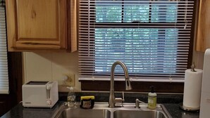 Une cuisine avec des meubles de rangement en bois, un évier à deux bacs, un grille-pain et une fenêtre avec des stores.