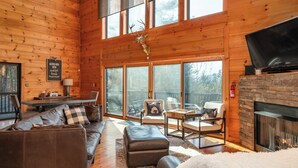 Interior acogedor de una cabaña con chimenea, mobiliario de cuero y ventanas amplias que ofrecen vistas al exterior.