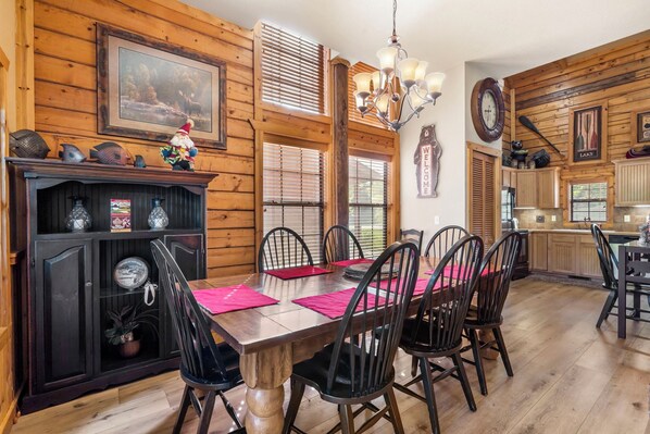 A wooden interior with a dining table, chairs, a chandelier, and a kitchen area.