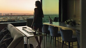 A modern interior with a balcony overlooking a cityscape at sunset, a dining area with chairs, and a kitchen counter with a bottle. - The Sun Rose West Hollywood (West Hollywood)
