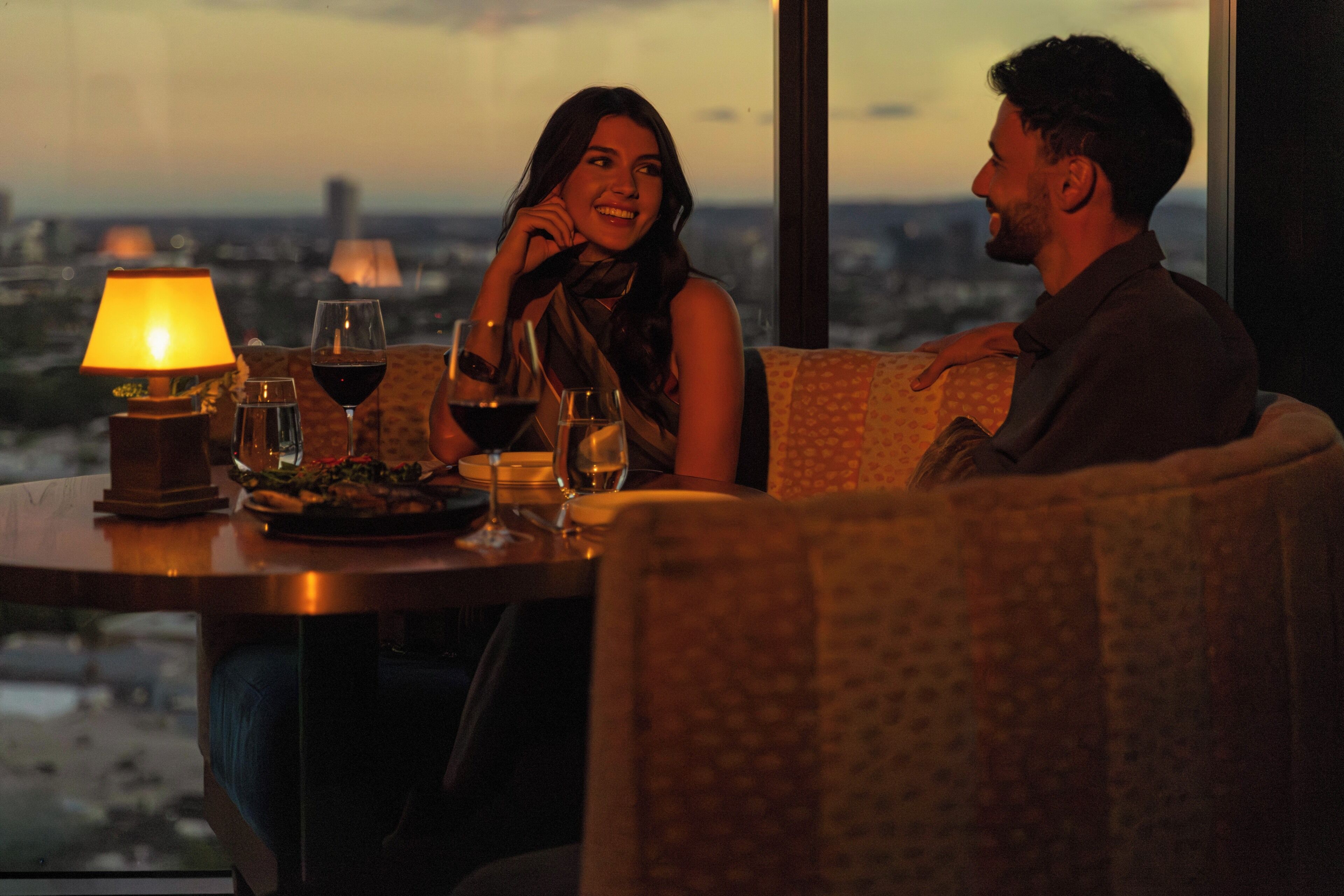 A couple seated at a table with a city view, a lamp, and wine glasses.