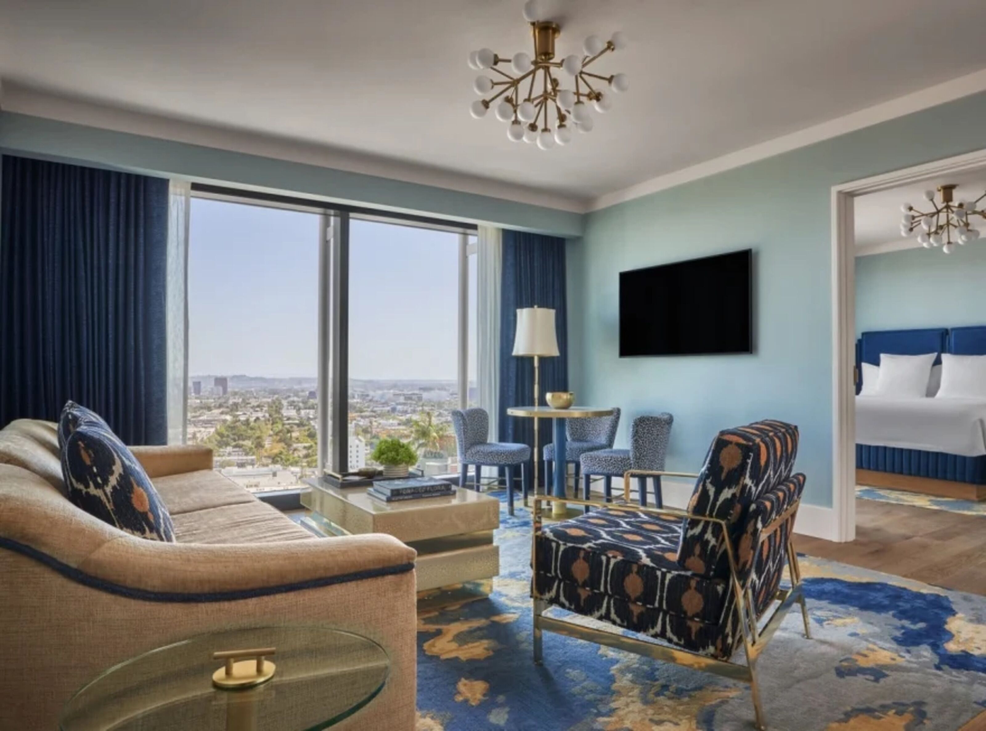 A hotel room with a large window, a sofa, a patterned armchair, a small table, and a bed with a blue headboard.