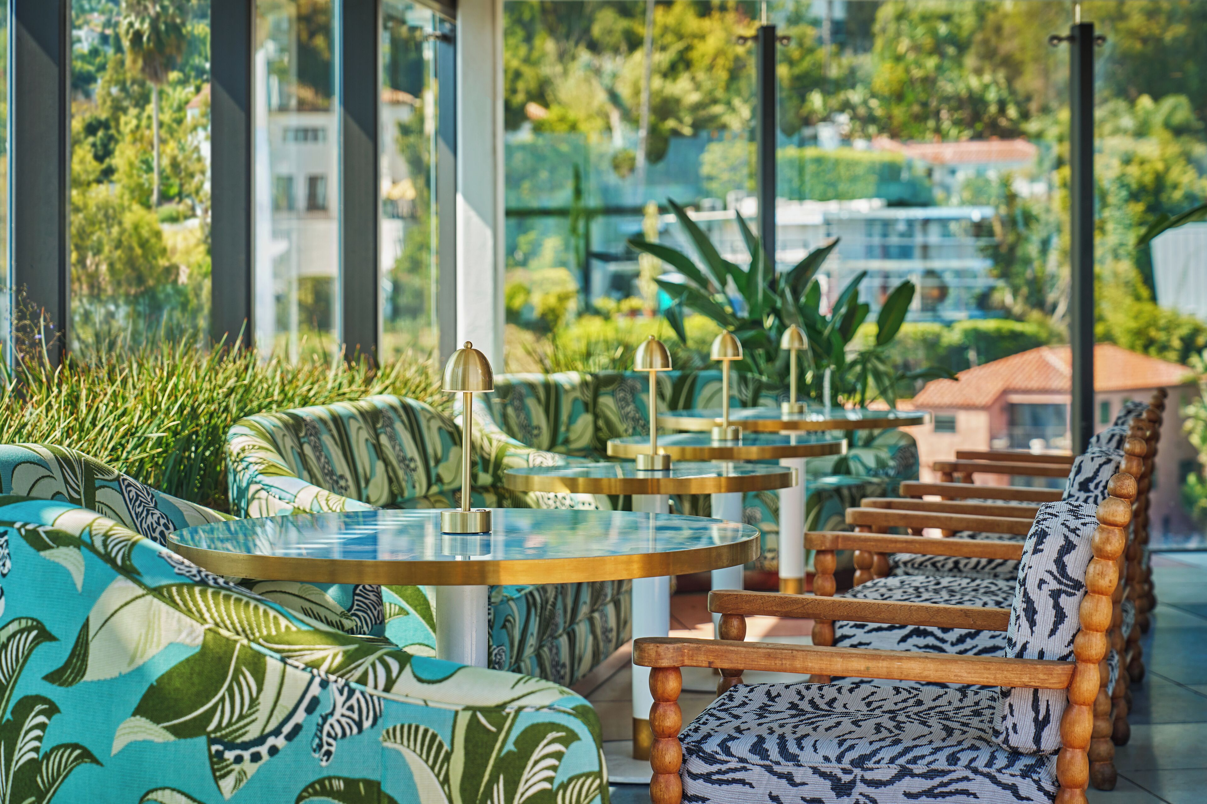 A row of wooden chairs and tables with patterned upholstery in a covered outdoor area with glass panels, surrounded by greenery and buildings.