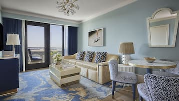 A modern hotel room with a sofa, armchairs, a coffee table, and a dining table. There is a large window with a view of the city.
