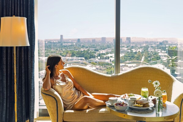 A person sitting on a sofa with a table of breakfast items in front of a large window overlooking a cityscape. - The Sun Rose West Hollywood (West Hollywood)
