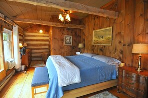 A wooden cabin bedroom with a bed, a bench, a dresser, a fan, a lamp, and a painting on the wall.
