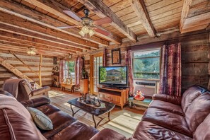 A log cabin interior with a television, leather sofas, a wooden coffee table, and a ceiling fan.