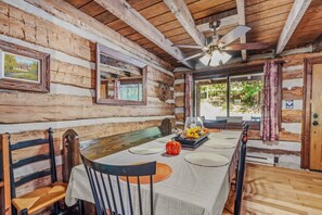 A log cabin interior with a dining table set for four, wooden benches, a ceiling fan, and a window with curtains. - Pondview Cabin - pond, log cabin, wifi, screen porch, deck, hiking, king bed (Berkeley Springs)