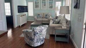 Una sala de estar con un sofá beige, un sillón estampado, una mesa de centro y un televisor sobre un mueble blanco.