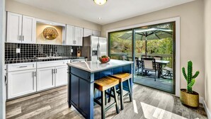 Una cocina moderna con armarios blancos, una isla azul y un amplio ventanal corredera que da a un patio exterior.