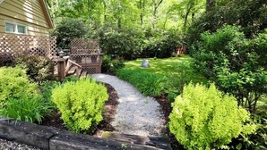 A house with a garden path leading to a wooden deck, surrounded by lush greenery. - Idlewood Cottage: Cozy Home w/ Covered Porch, Firepit, Yard | 8 Min to Downtown (Highlands)