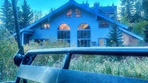 A blue house with a gabled roof and large windows, surrounded by trees. - True Slopeside 7-Bedroom Home: 75ft to Chair, Private Hot Tub, Deck, Sleeps 16 (Whitefish)