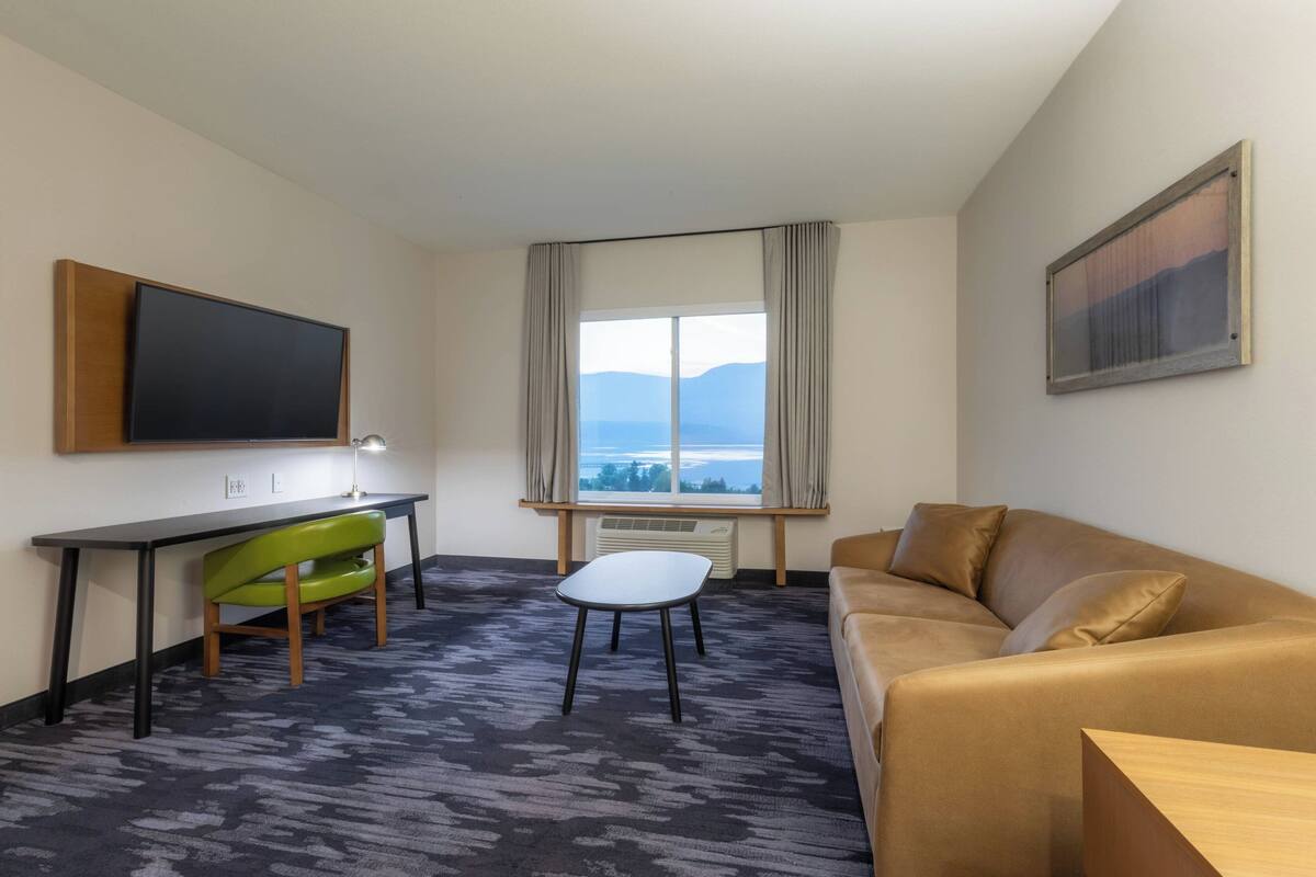 A hotel room with a flat-screen TV, a desk, a green chair, a white stool, a brown sofa, and a large window with a view of mountains.