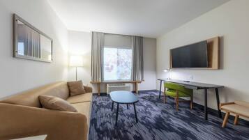 A hotel room with a sofa, a small table, a desk with a chair, a flat-screen TV, a window with curtains, and a radiator.