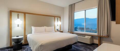 A hotel room with a large bed, two bedside lamps, a window with curtains, and a view of mountains.