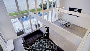 Ein geräumiges Zimmer mit großem Fenster zum Pool und Meer, eine gemütliche Sitzecke mit gemustertem Teppich sowie ein Essbereich mit modernen Stühlen und einem Glastisch.