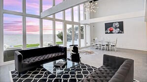 Ein geräumiges Wohnzimmer mit großen Fenstern, einem modernen Sofa, einem Glastisch und einem Essbereich mit weißen Stühlen.