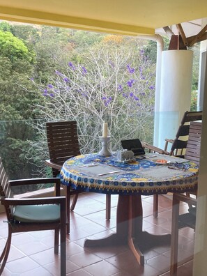 A round table with a floral tablecloth, surrounded by wooden chairs, set on a balcony with a view of trees and purple flowers.