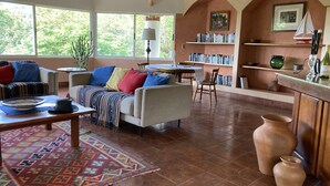 Un salón acogedor con un sofá, sillones, una mesa de centro y una alfombra estampada. Hay estanterías llenas de libros, una mesita con una lámpara y una ventana con vistas a zonas verdes.