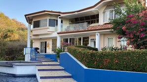 Una casa de dos pisos con piscina, escalones azules que conducen a la piscina y un jardín con flores rosadas.